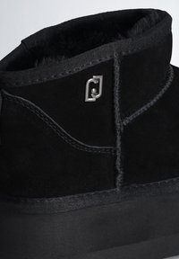 Stivale nero con tomaia in suede e una morbida fodera testurizzata. Include un logo metallico distintivo e dettagli di cucitura.