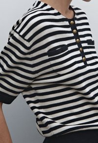 Randig stickad topp i svart och vitt, med rund halsringning, korta ärmar och fem guldknappar med fickor på bröstet.