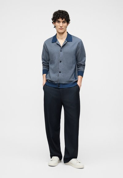 Νεαρός άνδρας στέκεται με τα χέρια στις τσέπες, φορώντας μπλε πουκάμισο με σχέδια και κουμπιά, σκούρο παντελόνι με λεπτές ρίγες και άσπρα αθλητικά παπούτσια, μπροστά σε απλό φόντο.