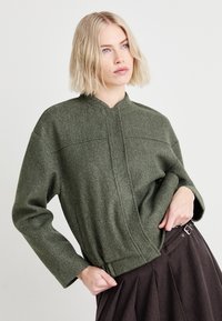 Maslinasto zelena, teksturirana jakna od vunenog materijala s uspravnim kragnom i skrivenim prednjim zatvaračem, s detaljima ušivenih panela i opuštenim krojem.