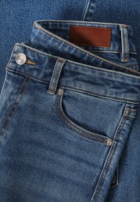 Jean en denim bleu à coupe droite, avec des accessoires métalliques argentés, des coutures orange contrastantes et une étiquette en cuir marron.