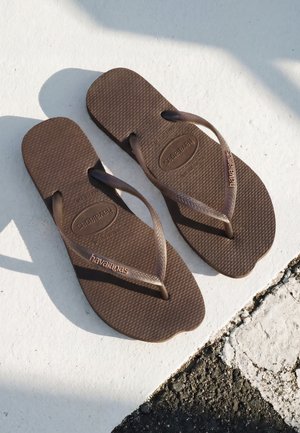 Coppia di infradito marroni Havaianas poggiati su una superficie bianca e nera testurizzata con la luce del sole che proietta ombre.