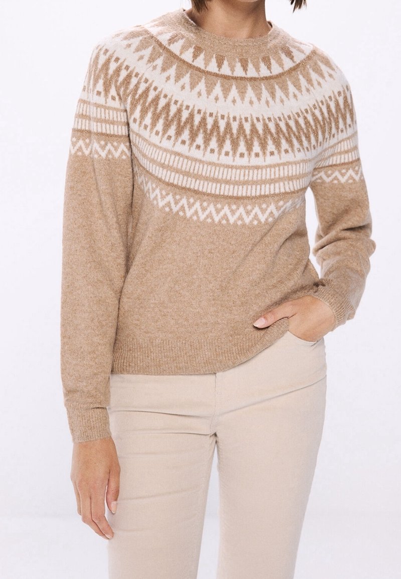 Beige Pullover mit weißen geometrischen Mustern über der Brust und den Schultern, getragen zu hellbeigen Hosen, Hand in der Tasche, neutraler Hintergrund.