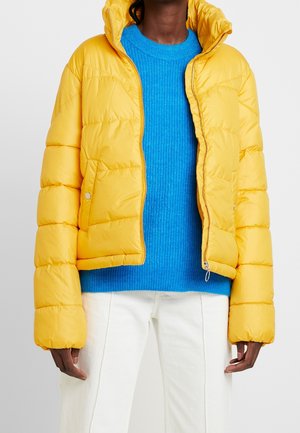 Personne portant une doudoune jaune vif sur un pull en maille bleu et un pantalon blanc, debout devant un fond clair uni.