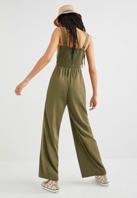 Olivgrön jumpsuit med vide ben, smockad byst och tjocka axelremmar. Bärs med en flerfärgad hatt och laced-up mönstrade skor.