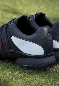 Pair of black and white golf shoes with spikes on green grass, shown from the back and slightly to the side.
