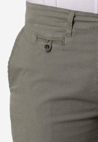 Primer plano de un bolsillo de pantalones verdes con una pequeña tapa asegurada por un solo botón marrón y negro.