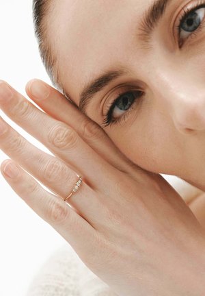 Anillo de oro con cuatro pequeñas piedras transparentes, llevado en una mano de piel clara, con los dedos tocando suavemente una mejilla. Fondo desenfocado.