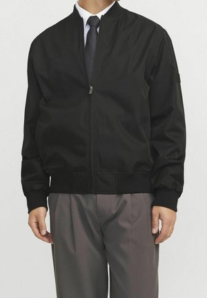 Personne portant une veste bomber noire zippée sur une chemise blanche et une cravate noire, associée à un pantalon gris plissé sur un fond uni.