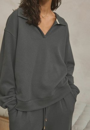 Woman wearing dark gray textured long-sleeve top with V-neck collar and matching pants, hands tucked in pockets, against neutral background.