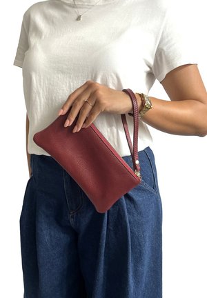 Borsa a mano in pelle rossa con finitura testurizzata, chiusura con zip e tracolla, appoggiata su una t-shirt bianca e pantaloni in denim.