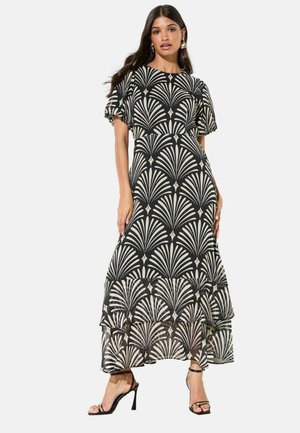 Nő fekete-fehér mintás maxiruhában, fodros szegéllyel és pöttyös ujjakkal, fekete magas sarkú szandállal párosítva.