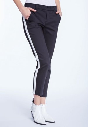 Persoon die zwarte slim-fit broek draagt met witte zijstrepen, handen in de zakken, gecombineerd met witte enkellaarzen.