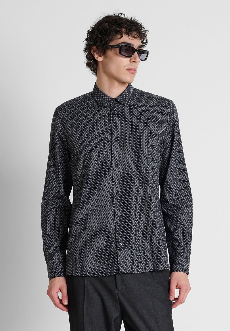 Hombre con camisa negra de manga larga con botones y estampado, pantalones oscuros y gafas de sol rectangulares negras mirando hacia un lado.