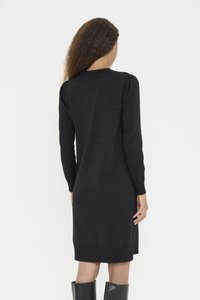 Göndör hajú nő hosszú ujjú fekete kötött ruhában és fekete térdig érő csizmában, háttal áll egy egyszerű fehér háttér előtt.