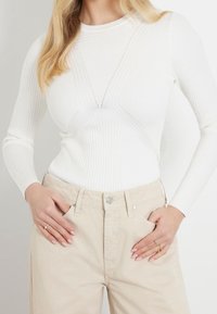 Weißes, geripptes Langarmoberteil mit Kreuzstichdesign, ergänzt durch beige, hoch taillierte Hose. Lässige Passform mit Rundhalsausschnitt und subtilen Details.