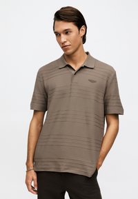 Άνδρας που φοράει καφέ πόλο μπλουζάκι με κοντά μανίκια και γιακά, μαύρο παντελόνι και ασημένιο βραχιόλι σε έναν καρπό.