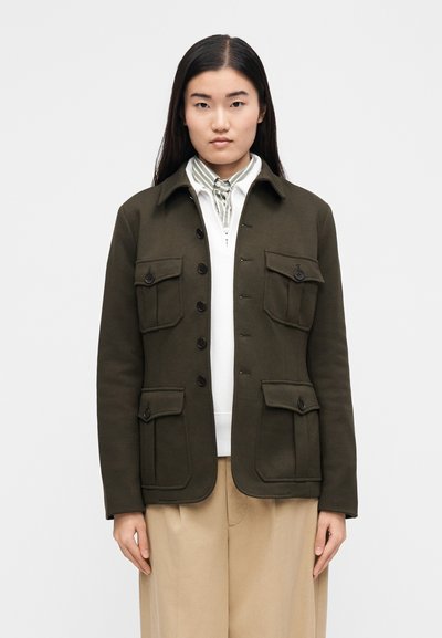 Olivno zelena jakna s štirimi sprednjimi žepi, zapiranjem na gumbe in ovratnikom; mehka tekstura s krojeno obliko. Nošena čez belo bluzo.