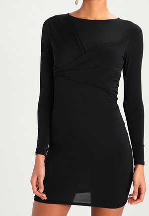 Kobieta ubrana w dopasowaną czarną mini sukienkę z długim rękawem, z dekoltem w kształcie łódki oraz subtelnymi marszczeniami na wysokości klatki piersiowej.