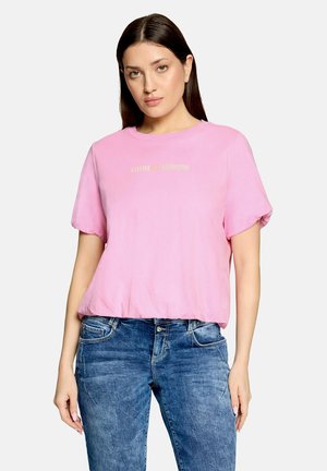 Woman with long dark hair wearing a light pink short-sleeve shirt with text and blue denim jeans, standing against a plain light background.