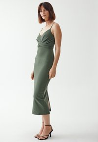 Grünes, ärmelloses Kleid mit verdrehtem Oberteil, knielangen seitlichen Schlitzern und einer figurbetonten Silhouette. Tragen mit schwarzen Riemchenschuhen.