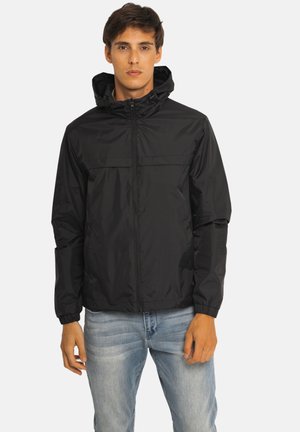 Jeune homme portant une veste à capuche noire zippée et un jean bleu clair, debout devant un fond uni clair.