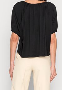 Sort løs top med rynket halsudskæring og korte, elastiske ærmer. Blødt stof former sig over kroppen og strækker sig til en buet kant.