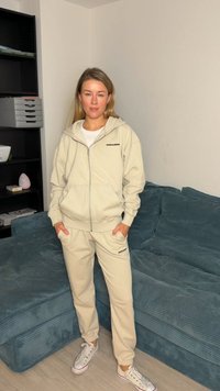Beige zip-up hoodie and matching joggers, both made of soft fabric, featuring a black logo on the chest and pants. White sneakers.