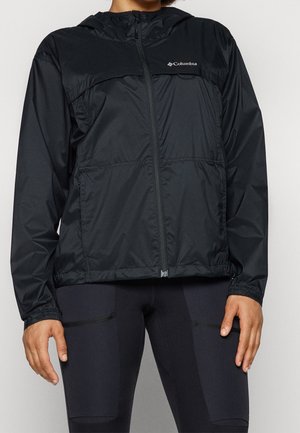 Person trägt eine schwarze Columbia Kapuzenjacke mit Reißverschluss und schwarze enganliegende Sportleggings, steht vor einem einfarbigen Hintergrund.