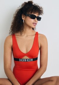 Červený plavky s hlubokým výstřihem do V a výřezem. Vyznačují se černým pasem s logem "CALVIN KLEIN". Nosené s černými obdélníkovými slunečními brýlemi.