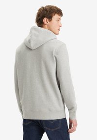 Sudadera gris con una textura suave y capucha con cordón. Presenta puños y dobladillo acanalados. Combinada con jeans azul oscuro. Vista trasera.