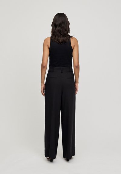 Calças largas pretas com cintura alta e passantes para cinto, apresentando uma textura suave e dois bolsos traseiros. Usadas com uma blusa de alças preta.