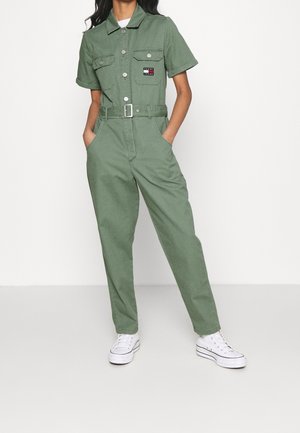 Personne portant une combinaison courte à manches courtes ceinturée de couleur vert olive avec des poches avant et des baskets montantes blanches, debout les mains dans les poches.