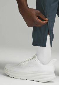 Spodnie sportowe w ciemnoszarym kolorze, z bocznymi zamkami na dole. Białe sneakersy z teksturowanym wierzchem i grubą podeszwą. Widoczna biała skarpeta do kostek.