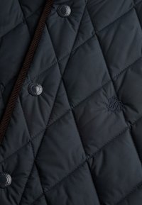 Quiltet marineblå jakke med diamantmønster, prydet med mørk kordfløjlskant, blanke sorte knapper og et broderet logo.