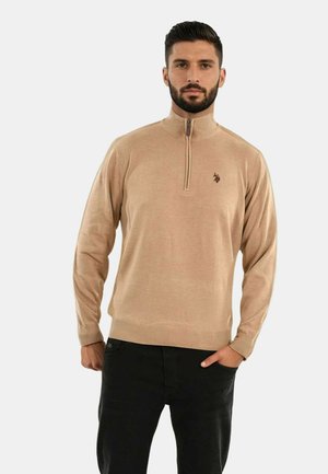 Beige Zip-Pullover mit hohem Kragen und gerippten Bündchen. Verfügt über ein kleines, dunkles Logo auf der Brust. Kombiniert mit schwarzen Denim-Hosen.