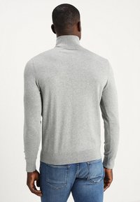 Uomo che indossa un maglione grigio chiaro a collo alto e jeans blu, di spalle su uno sfondo bianco uniforme.