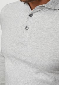 Primer plano de un hombre que lleva una camiseta polo de manga larga gris claro con dos botones negros abrochados cerca del cuello.