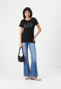 Zwarte T-shirt met het "GAP" logo in het grijs, gecombineerd met lichtblauwe flared jeans. Houdt een zwart studs handtas vast. Eenvoudige schoenen.