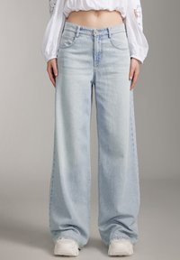 Jeans de mezclilla de pierna ancha en azul claro con una cintura alta, que cuentan con un cierre de botón y cremallera, bolsillos frontales y un sutil patrón desgastado.