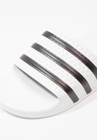 White slide sandal with three black stripes. Made of smooth rubber, featuring a textured strap and a flexible sole.