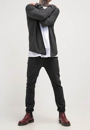 Persona con un cardigan gris oscuro, camisa blanca, pantalones negros y botas rojas hasta el tobillo, de pie con los brazos cruzados sobre el pecho frente a un fondo sencillo.