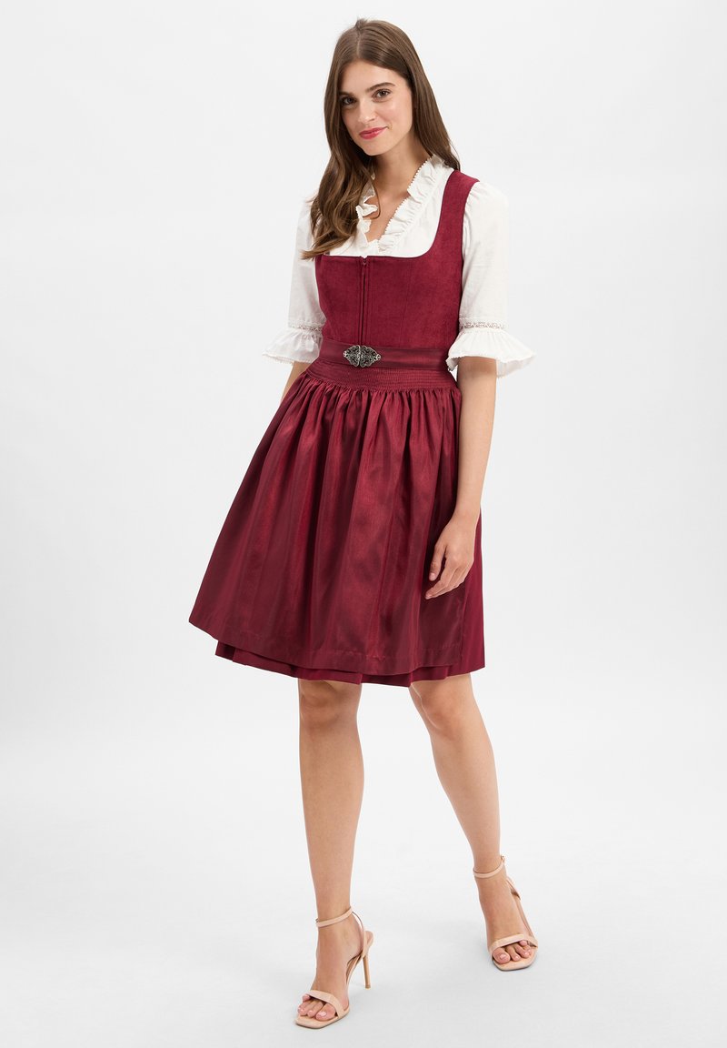Red dirndl dress with a fitted bodice, flared skirt, and ruffled white blouse. Features a decorative silver hardware accent at the waist.