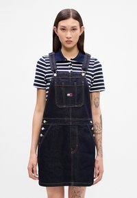 Tumši zili džinsu kopšārdzības kleita ar gaišiem diegiem, regulējamām siksnām, priekšējo kabatu un sānu pogām; valkāta virs tumši zila un baltā svītraina polo krekla.