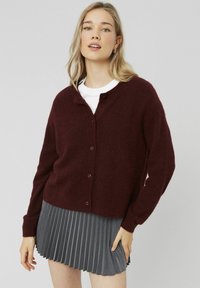 Gilet en laine bordeaux avec un col rond, des boutons sur le devant et des manches longues, associé à une jupe grise plissée. Texture lisse dans l'ensemble.