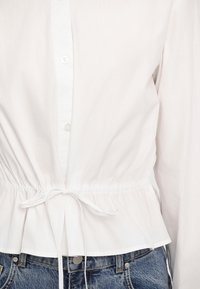 Blouse blanche boutonnée avec lien coulissant à la taille, associée à un jean en denim bleu, visible du milieu du torse jusqu’aux hanches.