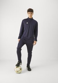 Marine-Trainingsanzug mit Viertelreißverschluss-Oberteil und passender Hose, mit reflektierendem Logo; Modell steht auf einem Fußball und trägt schwarze Fußballschuhe.