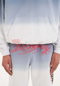 Sudadera y pantalones deportivos en degradado azul con desvanecido blanco. Cuenta con un bolsillo frontal, texto gráfico rojo y acentos florales en los pantalones.