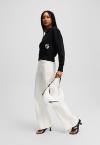 Μαύρο κοντό sweatshirt με λευκό λογότυπο, συνδυασμένο με λευκά παντελόνια με ψηλή μέση και φαρδιά πόδια. Κρατάει μια λευκή στρογγυλή τσάντα χειρός με λεπτομέρεια λογότυπου.