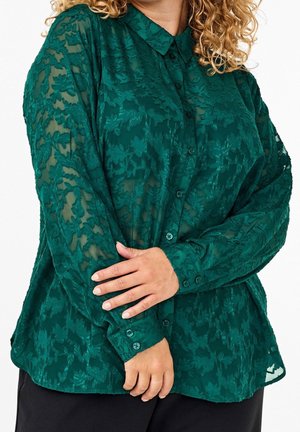 Kobieta o kręconych włosach, ubrana w zieloną przezroczystą bluzkę w kwiatowy wzór zapinaną na guziki oraz czarne spodnie, z założonymi na wysokości talii ramionami.